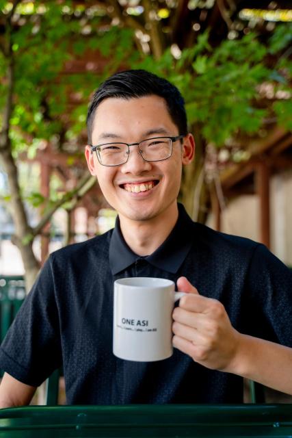 A picture of a man holding a mug underneath green foliage,